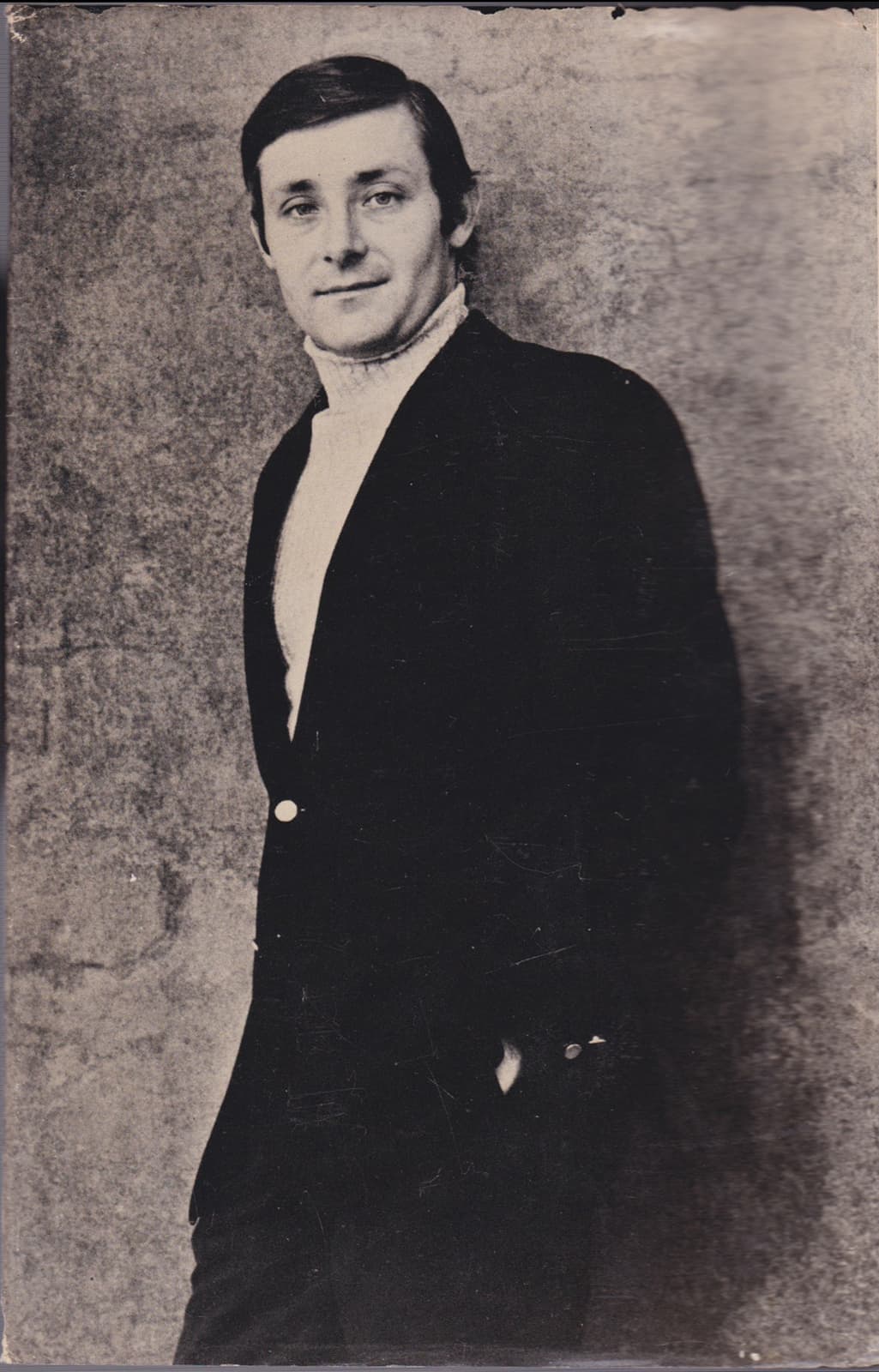 Peter Benchley in a dark jacket and white roll-neck, leaning against a mottled wall, hand in pocket
