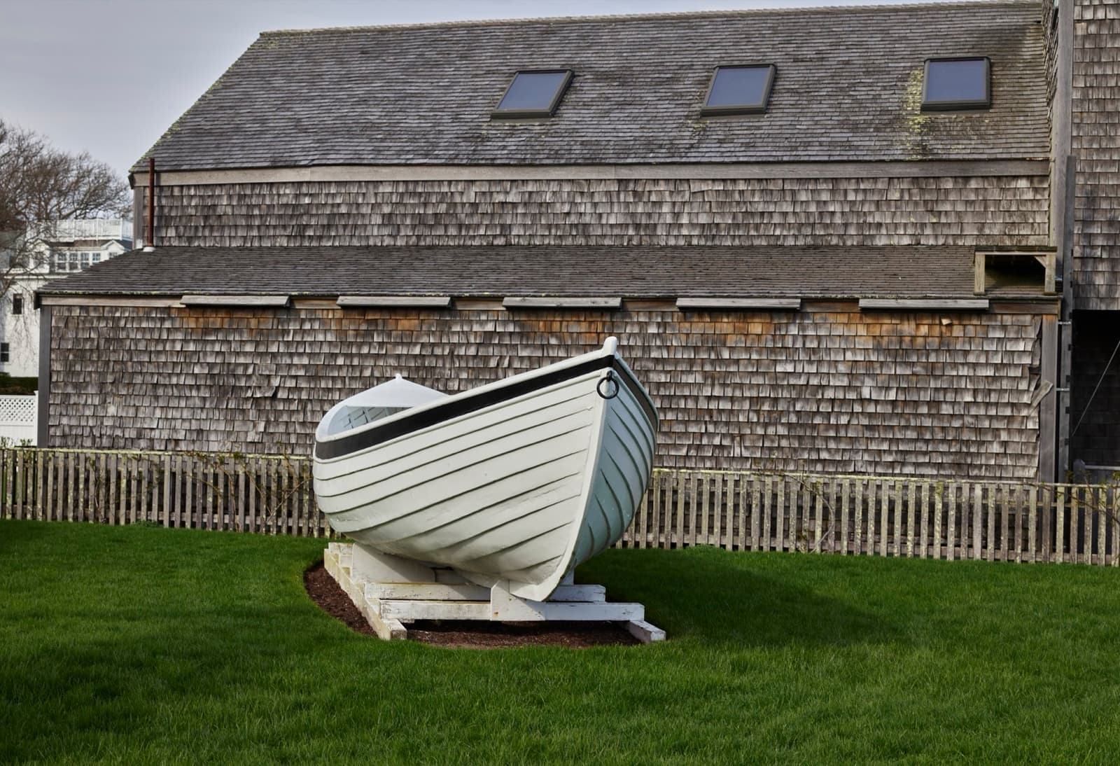 A weathered shingle boat shed with a white rowing boat on the lawn in front