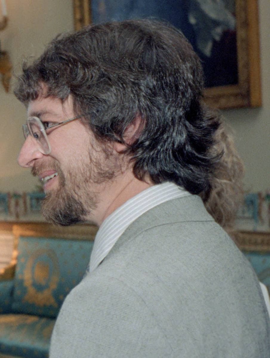 A bearded young Steven Spielberg in glasses and a grey jacket, smiling in a blue-walled room