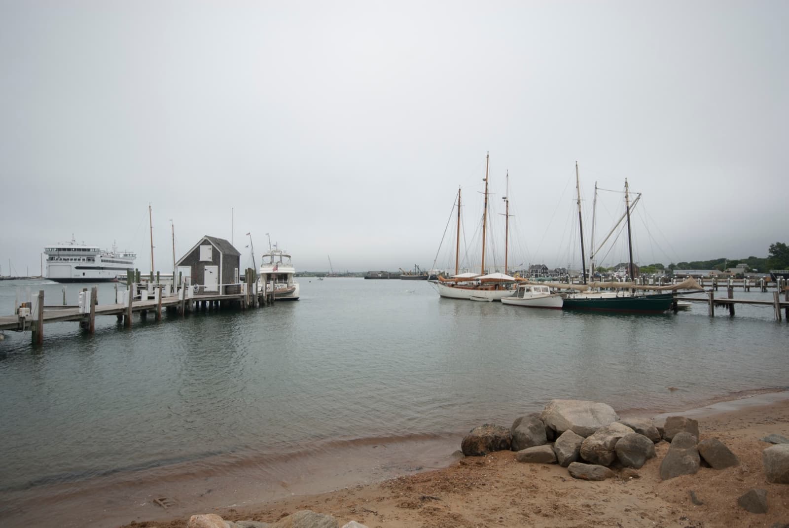 Grey harbour water with piers, a ferry and tall-masted schooners under a white sky