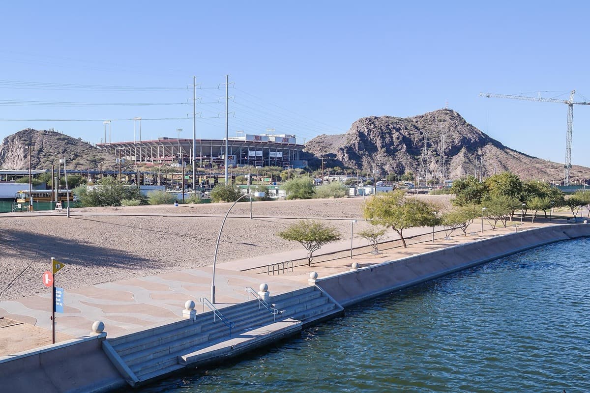 A stadium roofline beyond a desert park, a canal and rocky hills