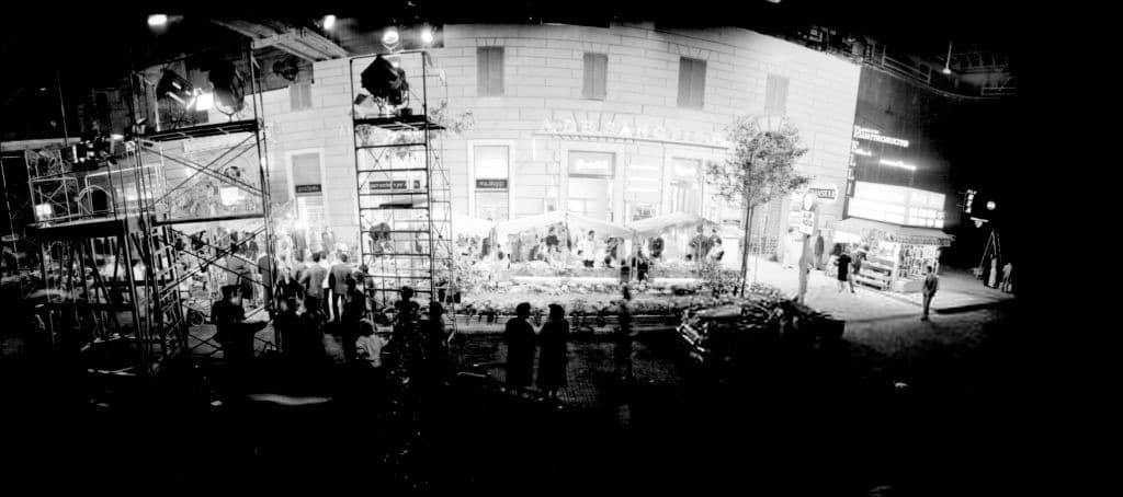 A night film set: a lit street of café awnings and hotel fronts under scaffolding and lamps