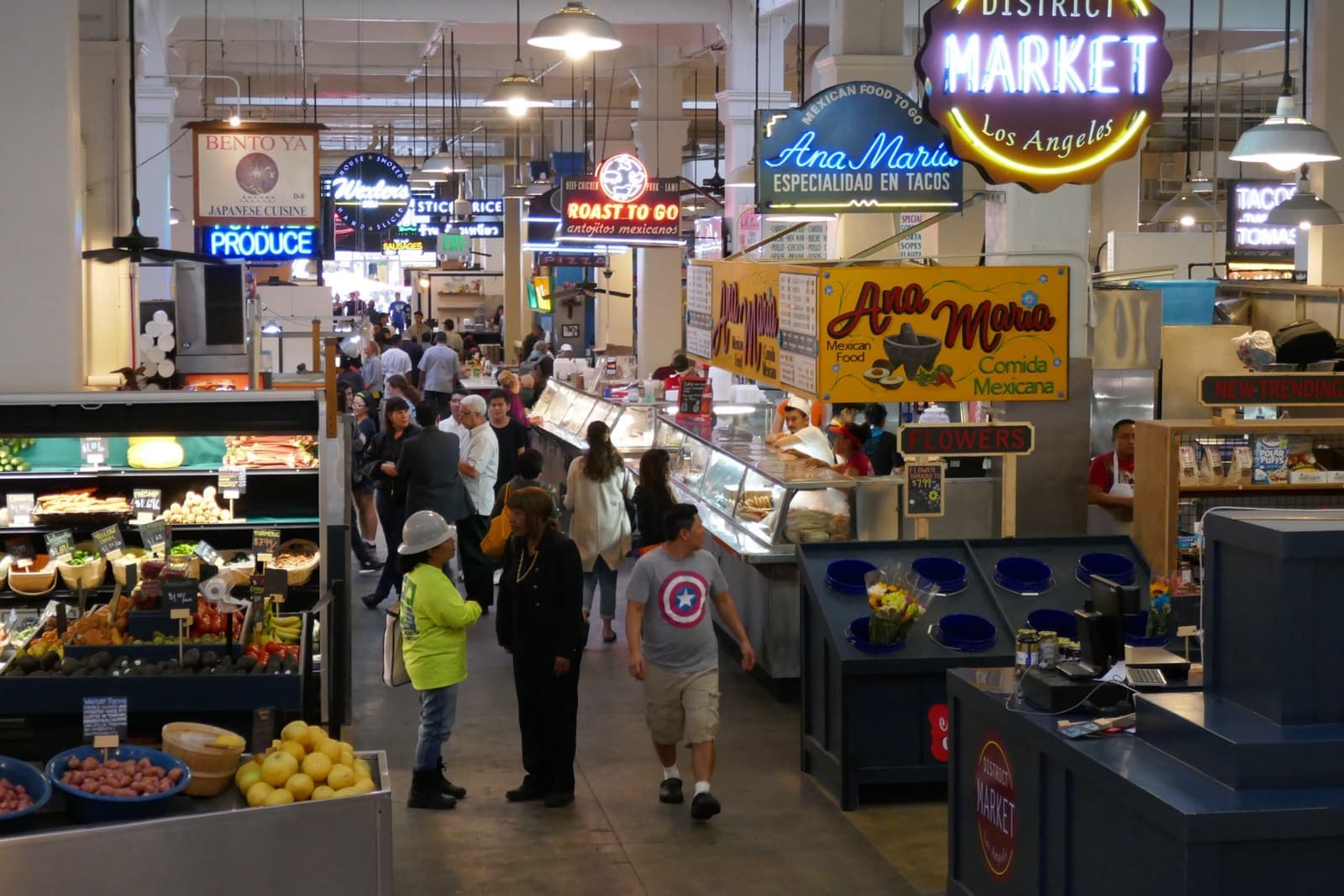 An aisle of Grand Central Market with neon stall signs, produce and shoppers