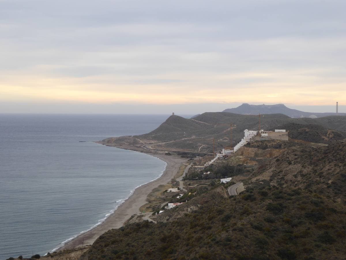 A long grey beach curving under brown hills beside a calm sea