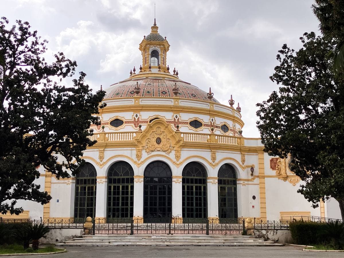 A white and ochre domed pavilion with arched windows behind palm trees