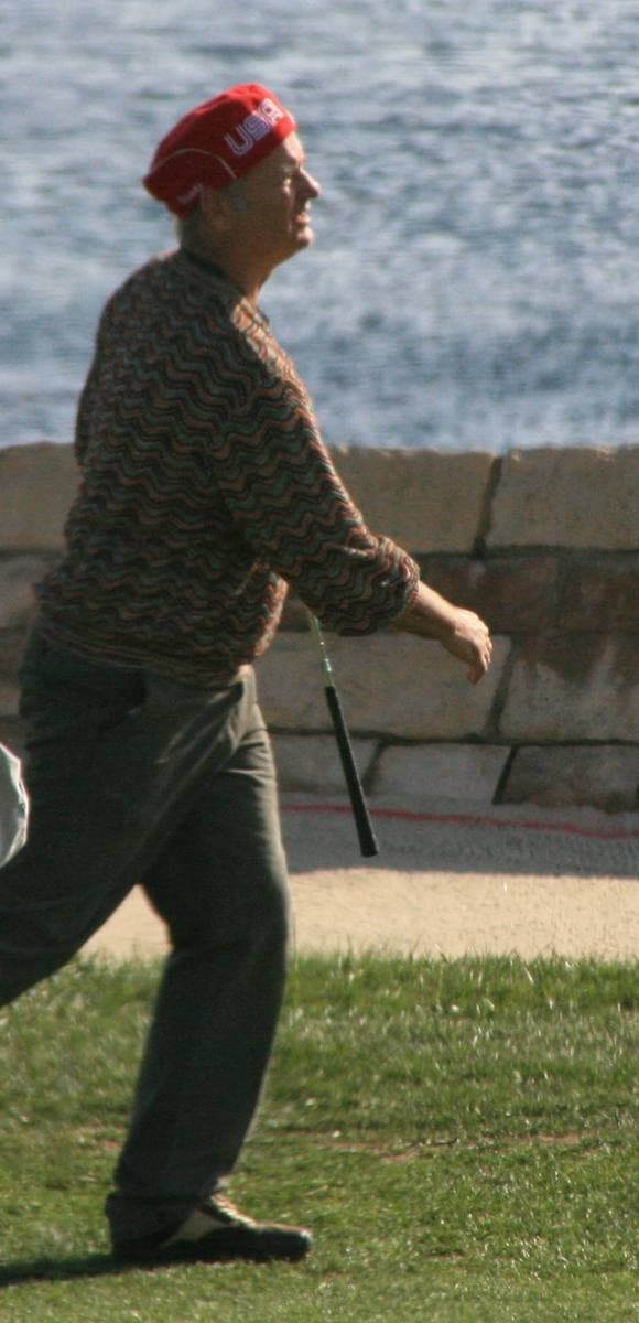 Bill Murray in a red cap and dark jumper striding across a golf green by the sea