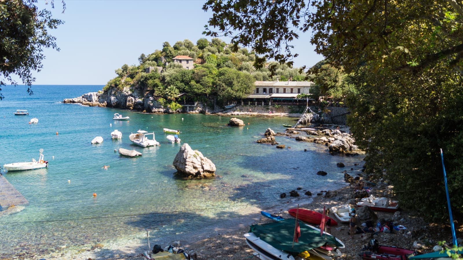 A small stone harbour with fishing boats, a big boulder in the water and a wooded headland