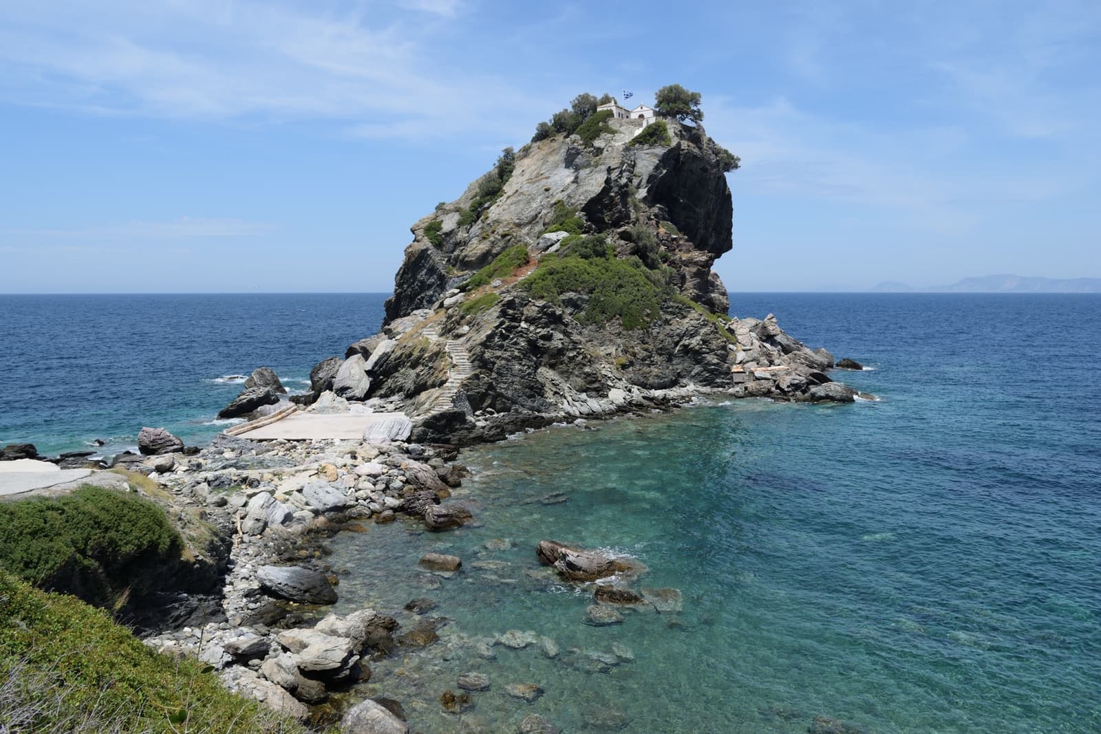A rocky headland with a chapel on top, a pebble shore and clear turquoise water in front