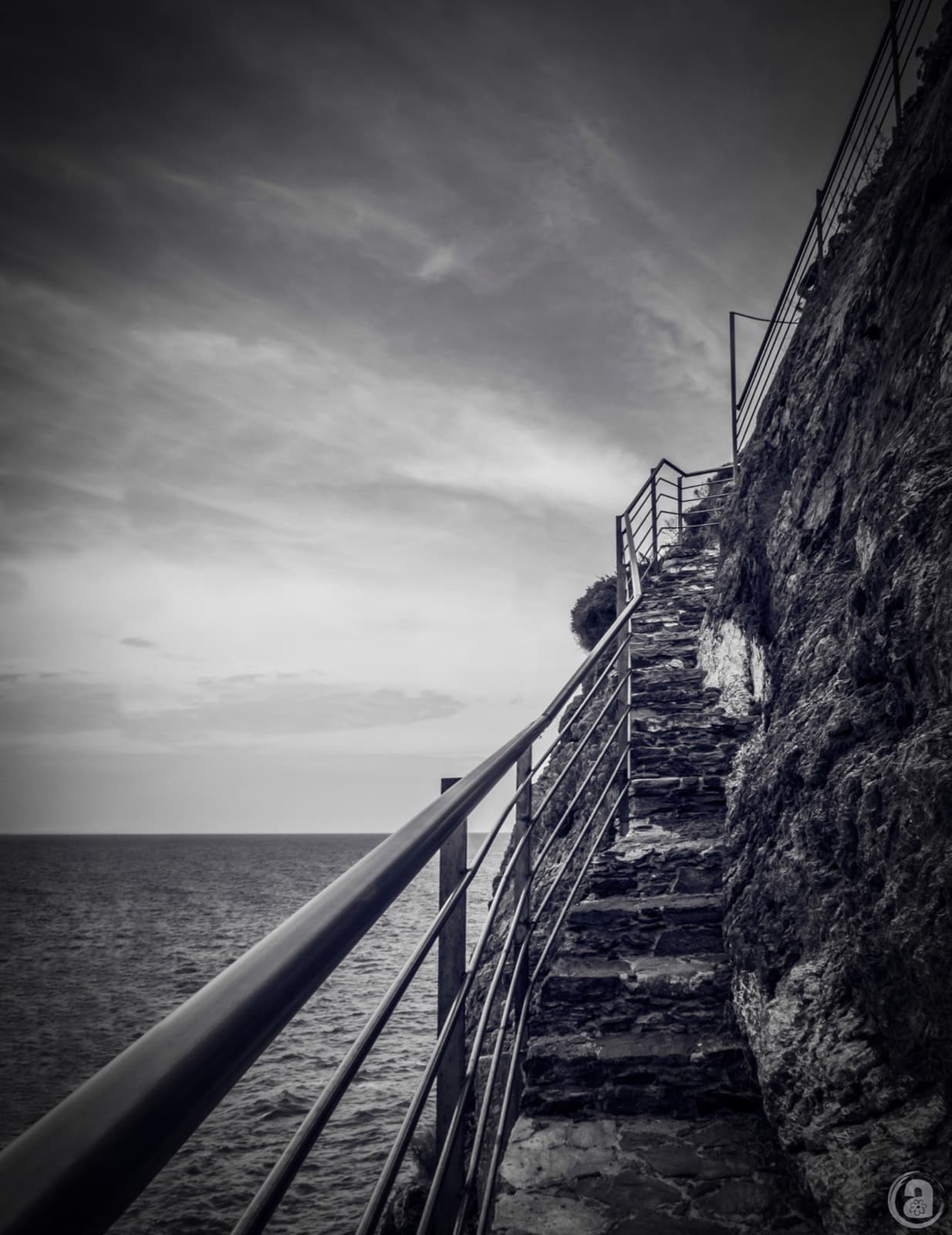 Black and white photograph of a narrow stone stair with a metal handrail climbing a cliff above the sea