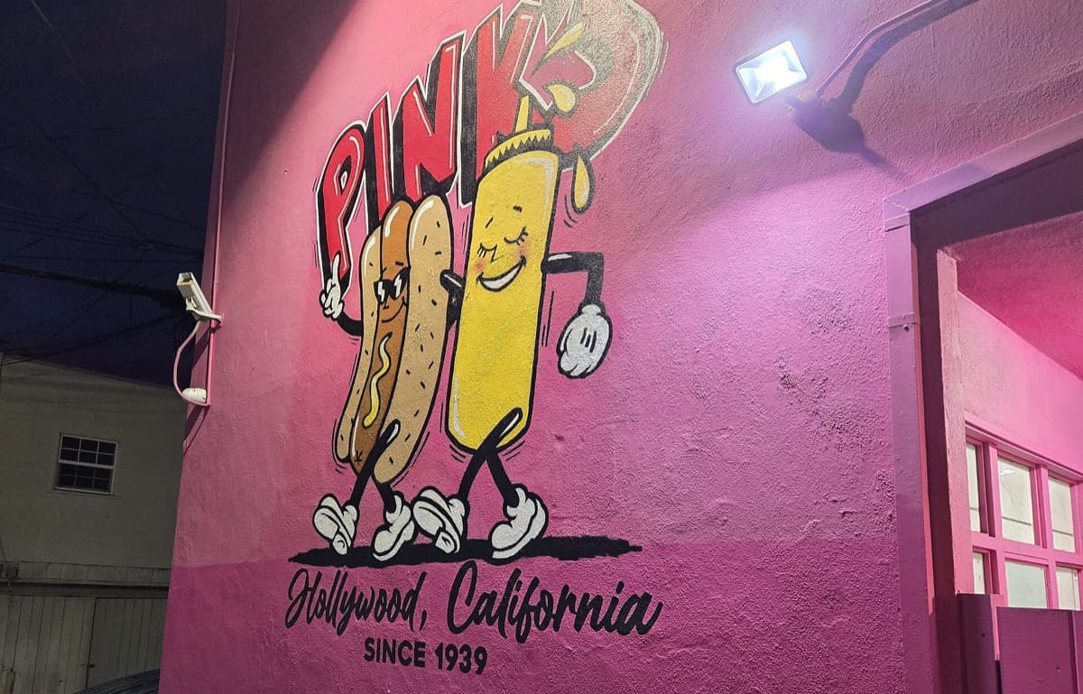 A small hot dog stand with a red sign and a queue on a busy Los Angeles avenue