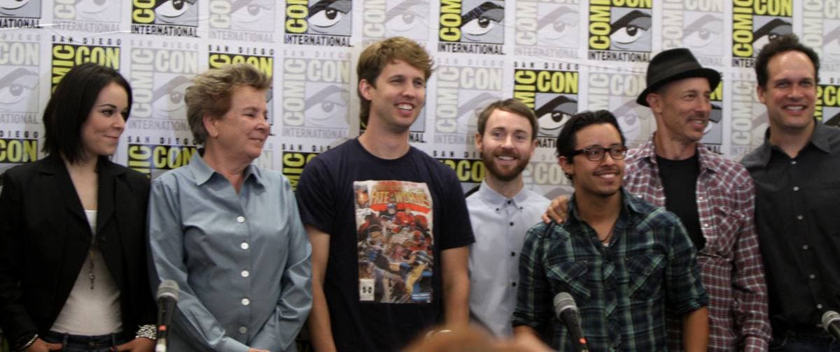 Seven members of the cast standing in a row in front of a Comic-Con backdrop