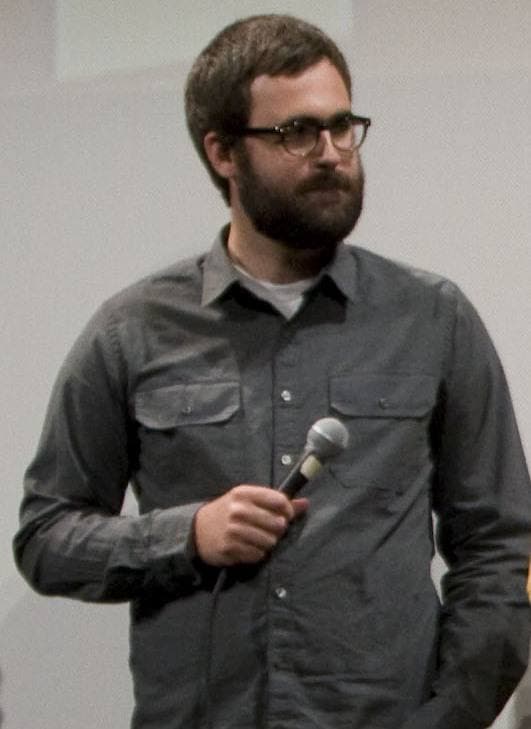 Jared Hess, bearded, in glasses and a grey shirt, holding a microphone