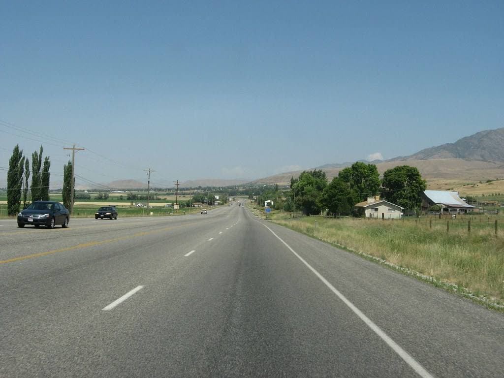 A straight two-lane highway running between fields towards low hills