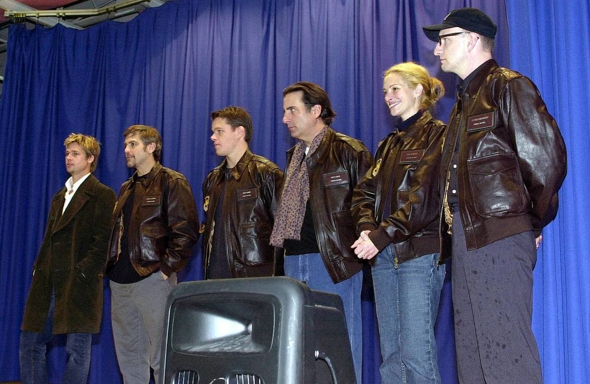 Six people in matching brown leather flight jackets standing in a row in front of a blue curtain