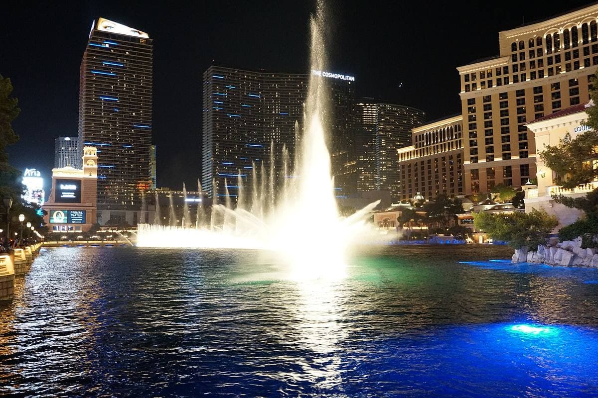 A tall white plume of water lit at night on a dark lake, hotel towers behind