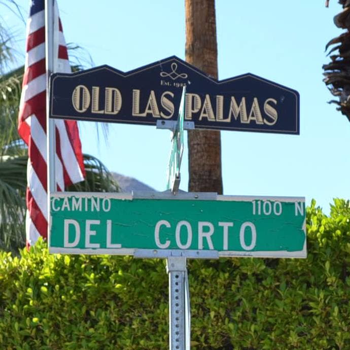 An OLD LAS PALMAS sign on a pole over a green street sign, palms behind