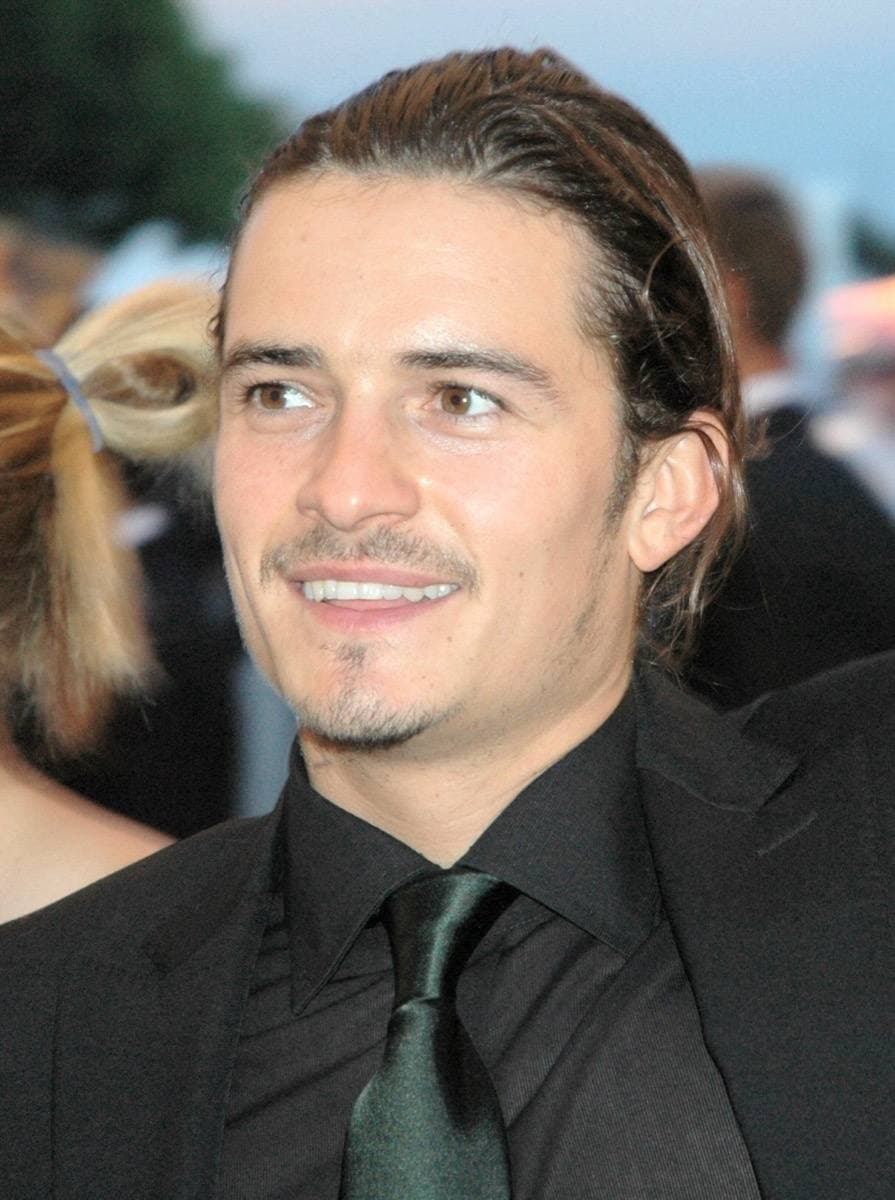 Orlando Bloom, hair swept back, in a dark shirt and green tie, smiling to one side