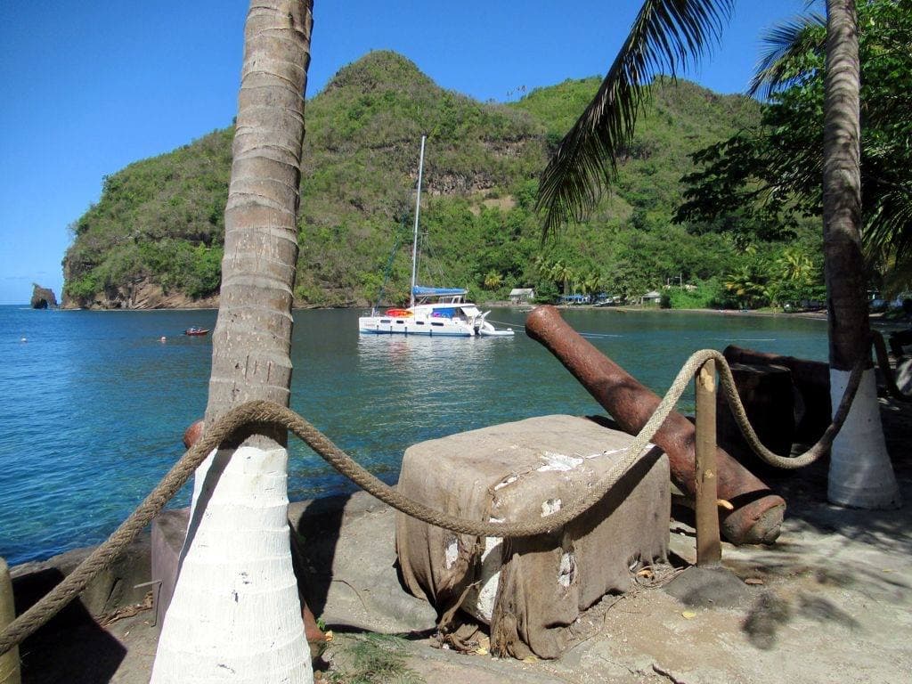 A rusted cannon beside a palm trunk wrapped in rope, a catamaran in the green bay beyond