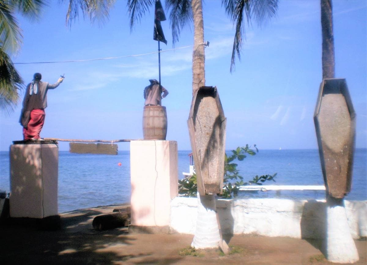 Two upright wooden coffins and a pirate figure on a whitewashed sea wall above blue water