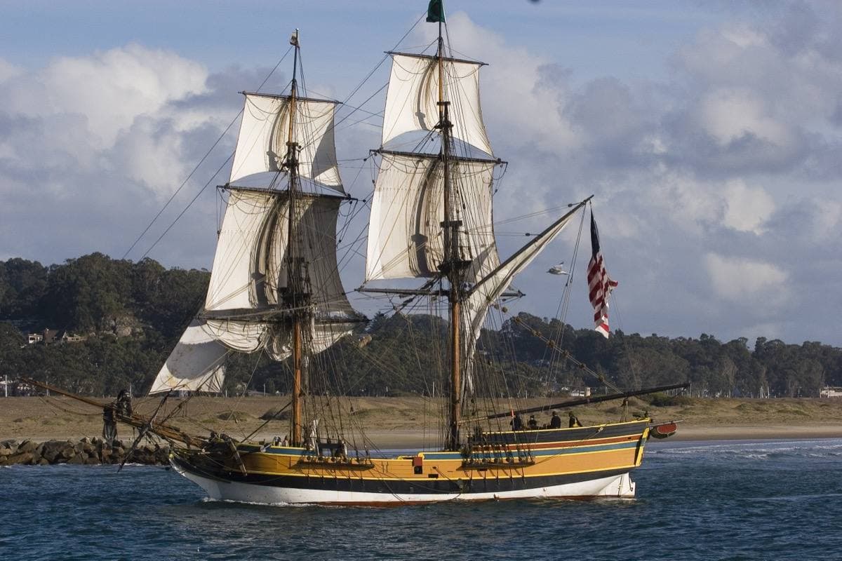 A two-masted brig with cream sails and a yellow and blue hull sailing across a bay