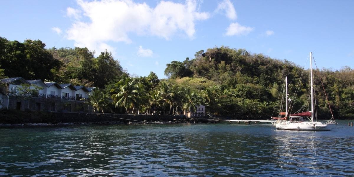 A green bay with a yacht at anchor and low waterfront buildings under wooded hills