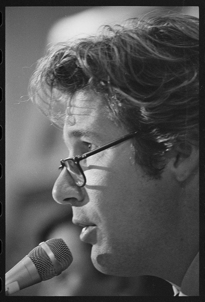 Black-and-white profile of Richard Gere in glasses speaking into a microphone