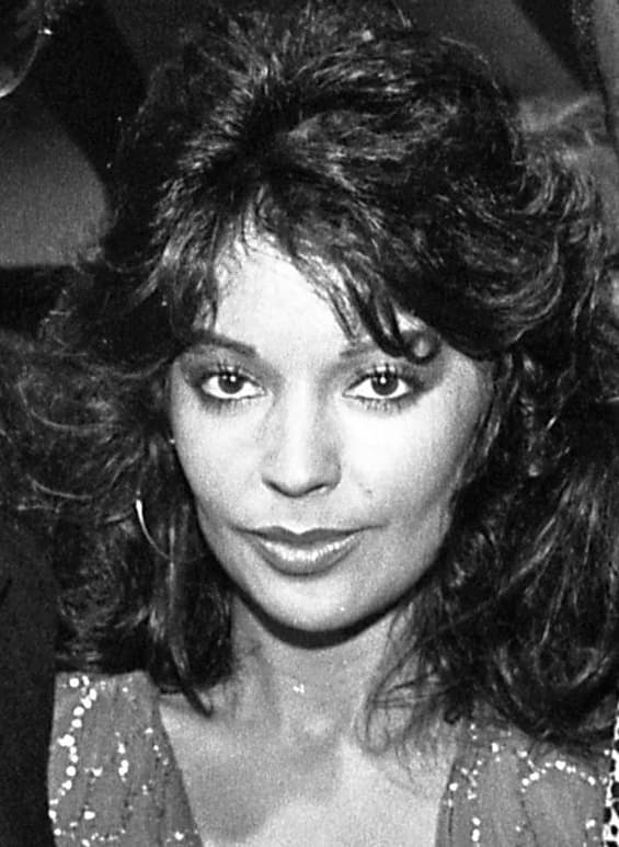A smiling young woman with long dark curled hair and hoop earrings, black and white