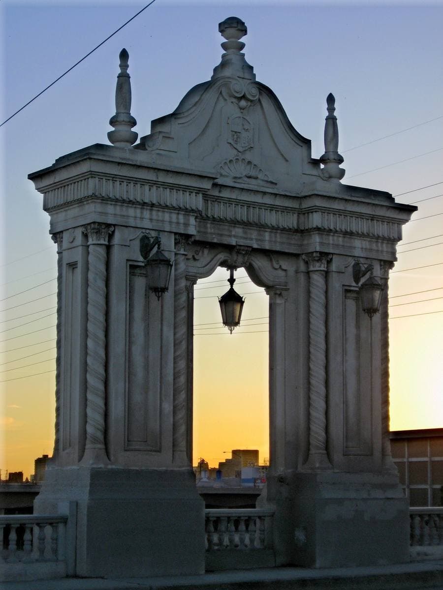 An ornate grey stone arch with a hanging lamp, against a dusk sky