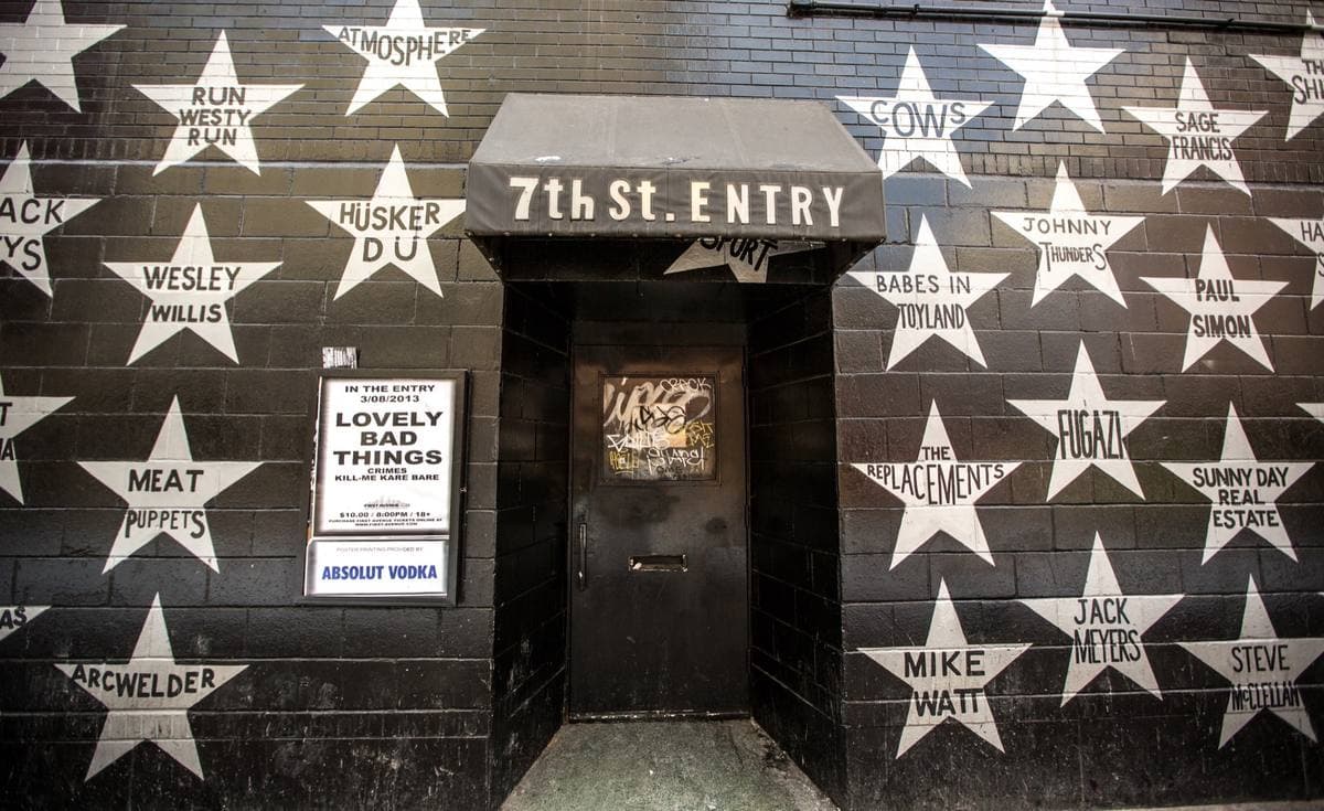 A black brick wall covered in painted silver stars with band names, around a doorway under a 7th St. Entry awning