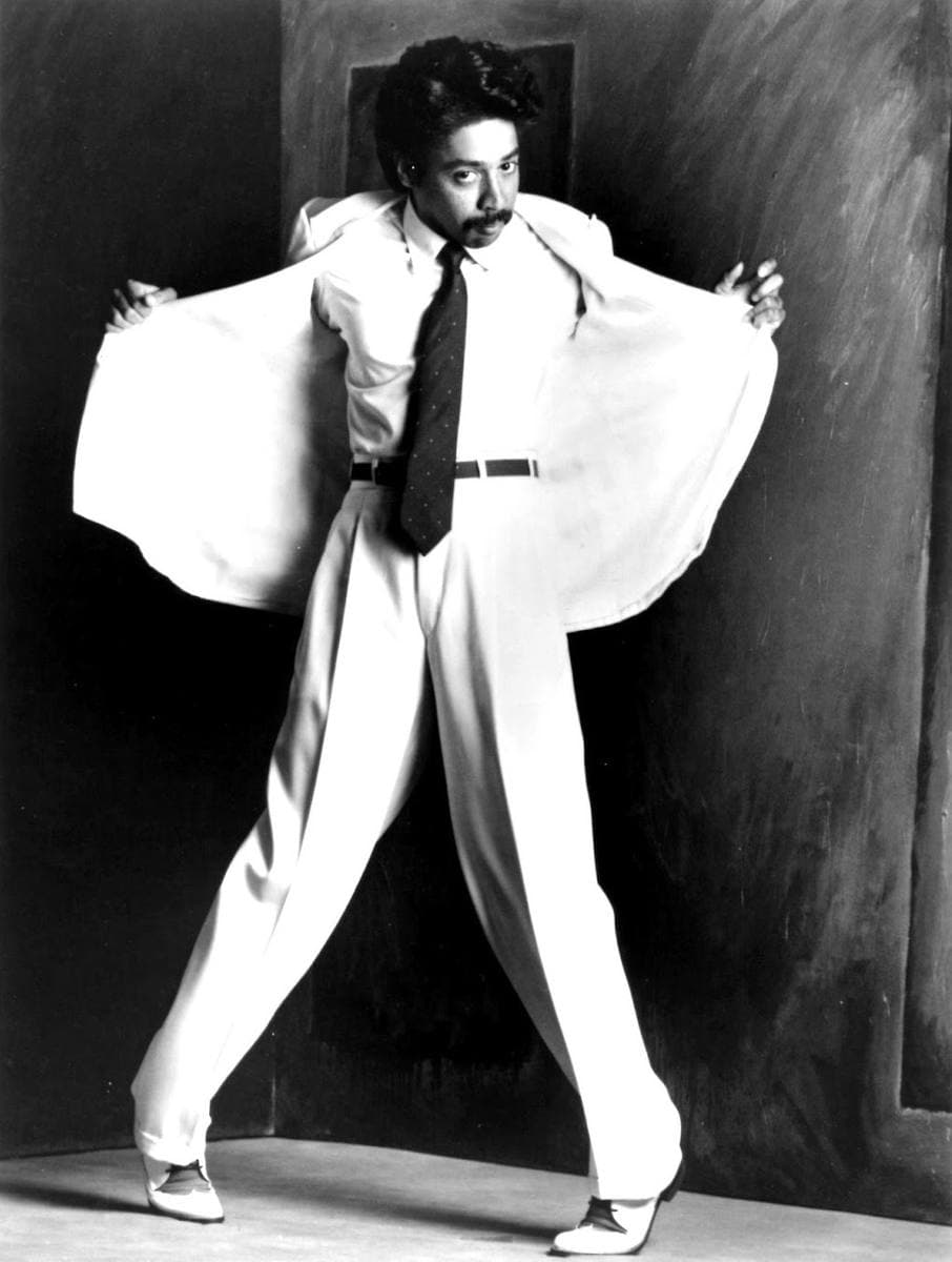 A man in a pale double-breasted suit and tie holding his jacket open, black and white