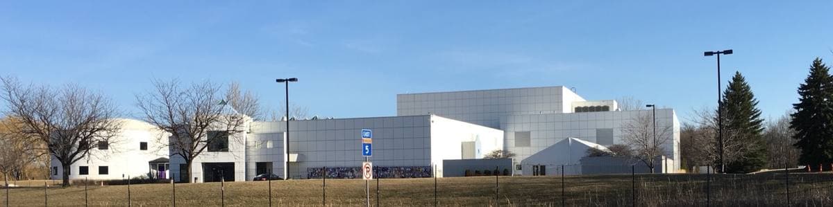 A long white and grey building complex behind a field and bare trees