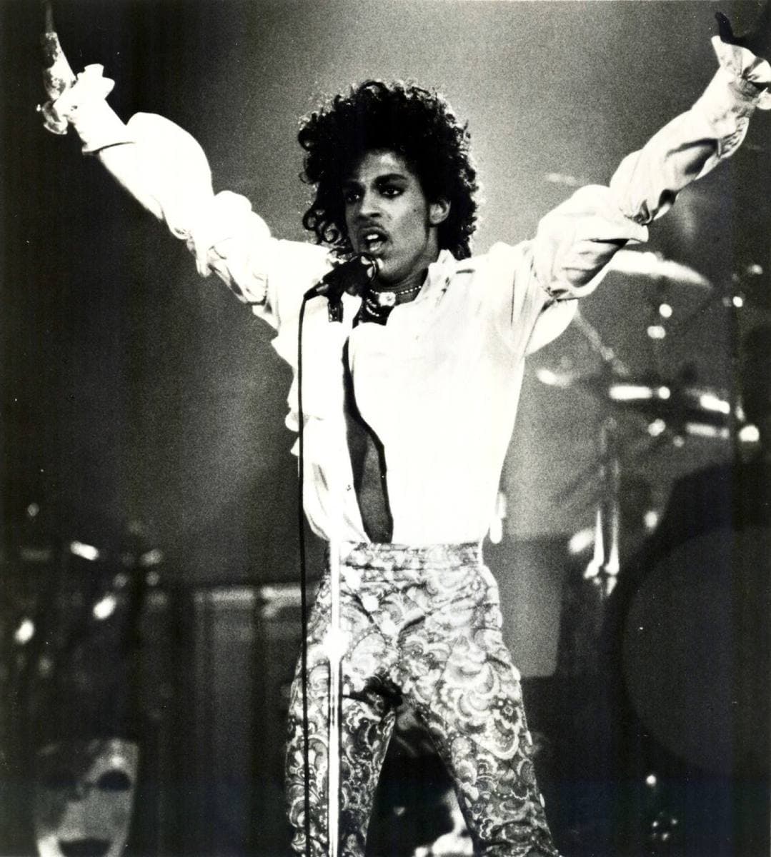 Prince on stage with both arms raised, in a ruffled white shirt and patterned trousers, black and white
