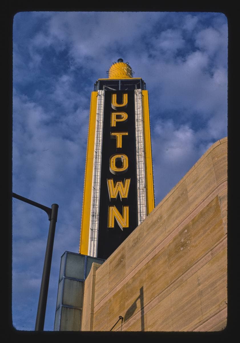 Colour photograph of a tall black and yellow sign tower spelling UPTOWN against a blue sky with clouds