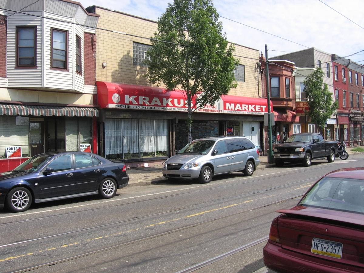 A row of shops on a sunny street with tram tracks, including a red-fronted Polish supermarket