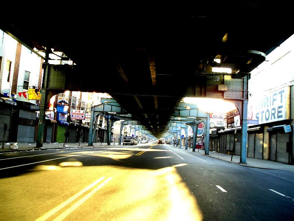 A sunlit avenue under the dark steel deck of an elevated railway, shopfronts on both sides