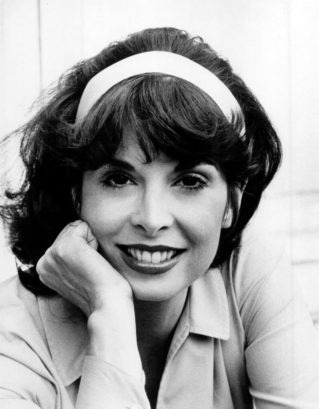 Talia Shire smiling, chin on her hand, wearing a white hairband