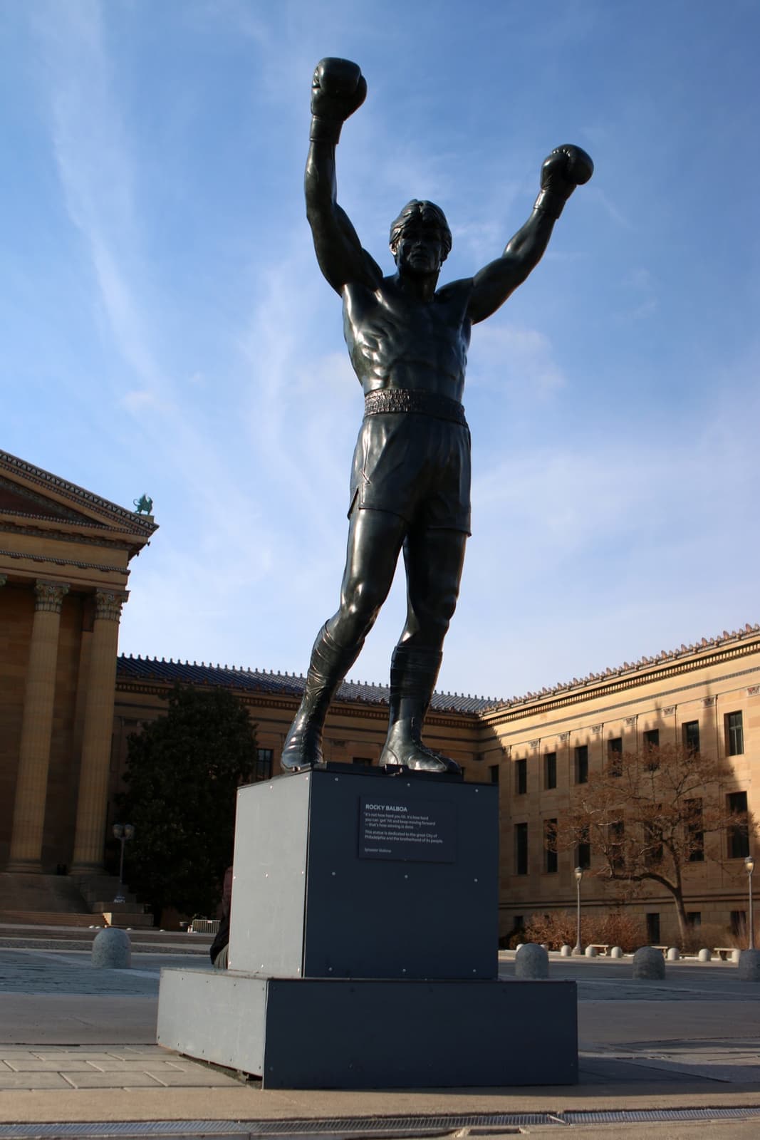 Bronze statue of Rocky Balboa with both gloved fists raised, on a plinth in front of the Philadelphia Museum of Art under a blue sky