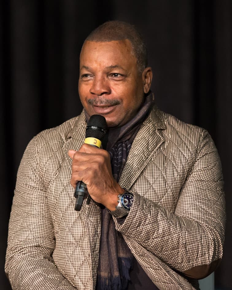 Carl Weathers speaking into a microphone, in a checked jacket and scarf