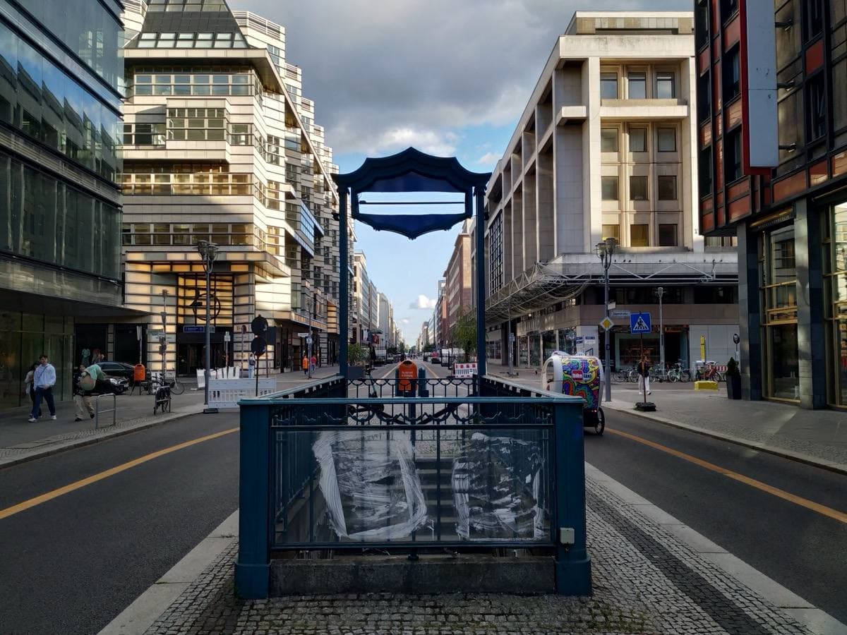 A fenced-off U-Bahn stair entrance with a blue station sign in the middle of a wide city street