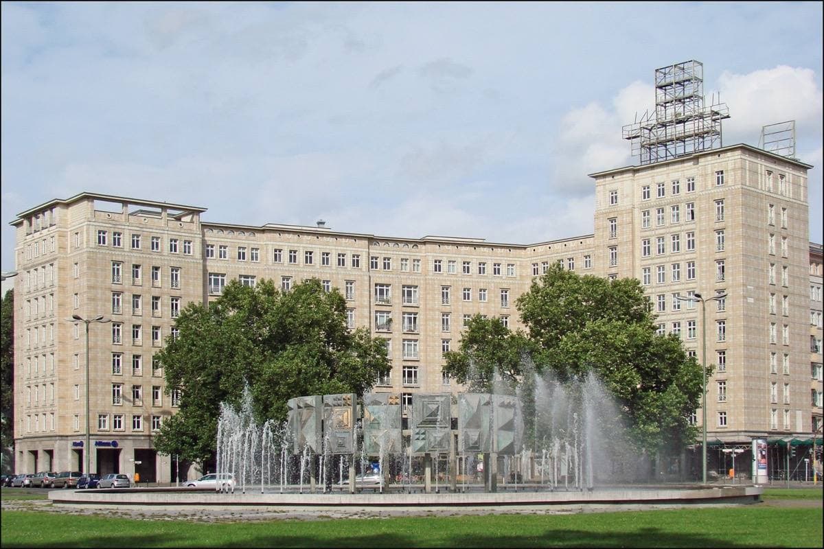 A fountain in front of tall pale apartment blocks crowned by a tower