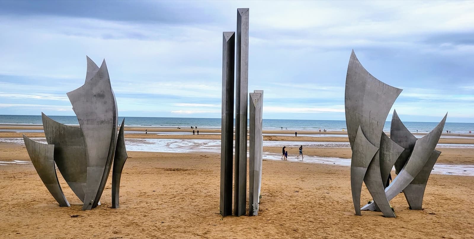 Tall stainless-steel sculpture forms standing on a wide beach at low tide