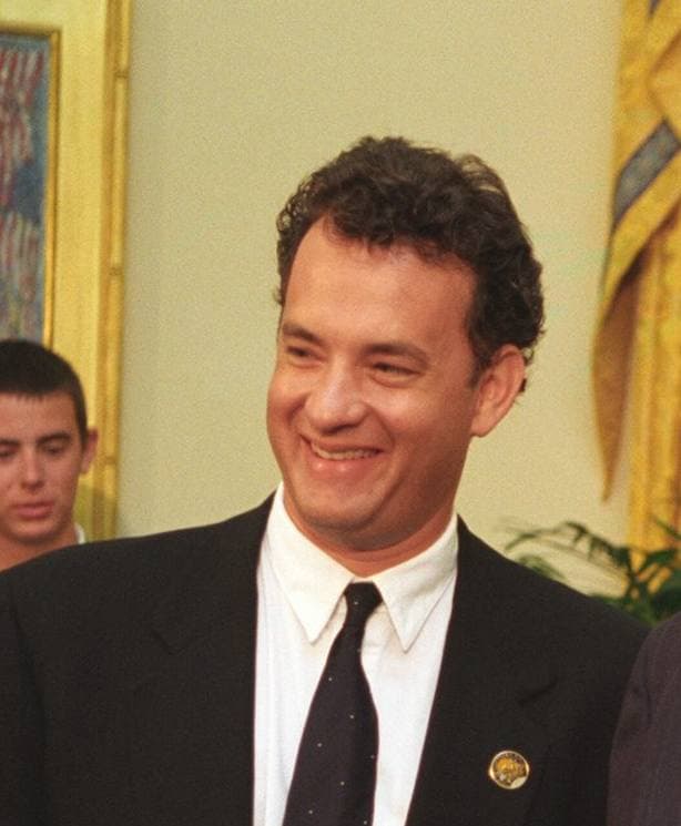 Tom Hanks, smiling, in a dark suit and tie in the Oval Office