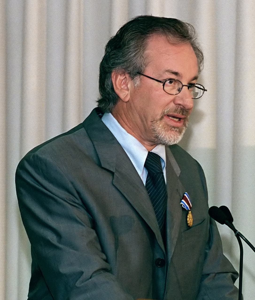 Steven Spielberg speaking at a lectern, a medal on his lapel