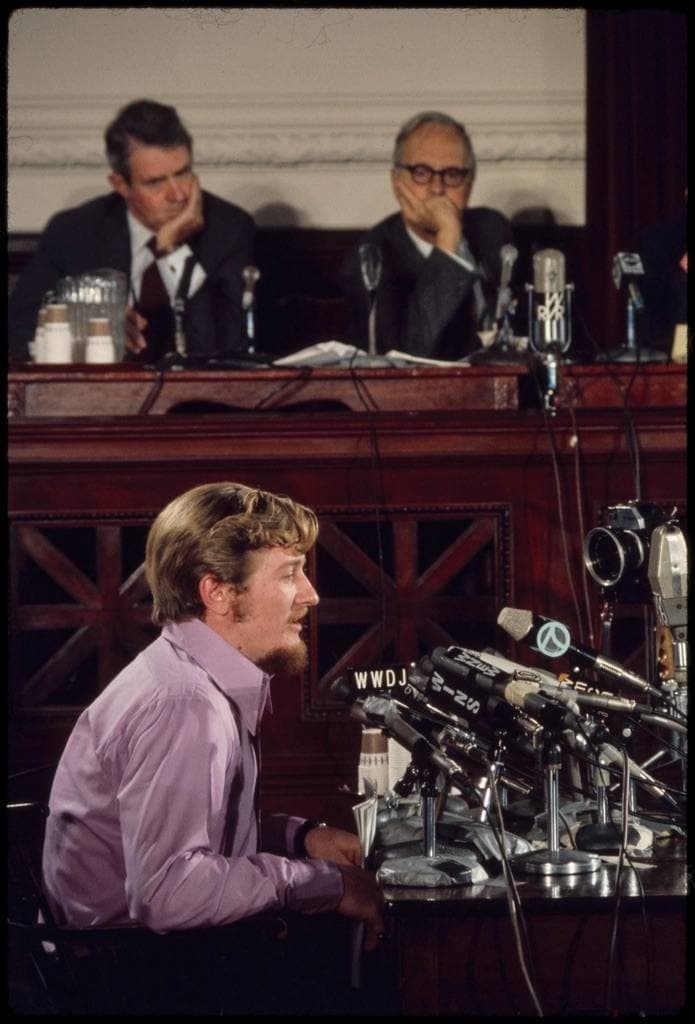 A young man in a lilac shirt at a table crowded with microphones, men at a panelled dais behind him
