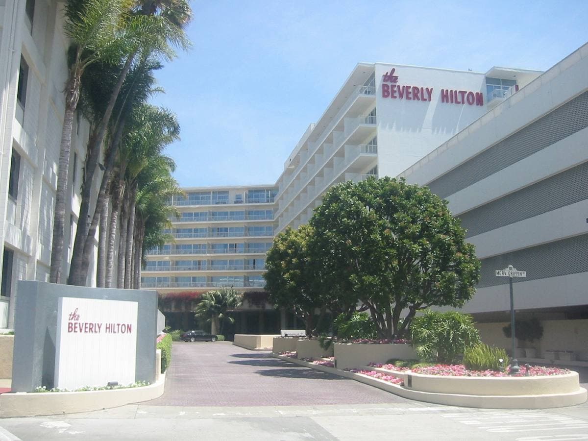 Colour photograph of a long white modernist hotel behind palm trees, with a sign reading The Beverly Hilton