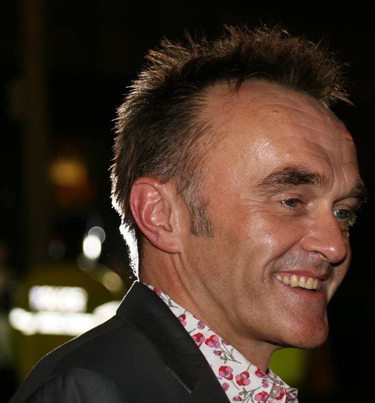 Colour photograph of a smiling man with spiky hair in a dark jacket and patterned shirt, at night