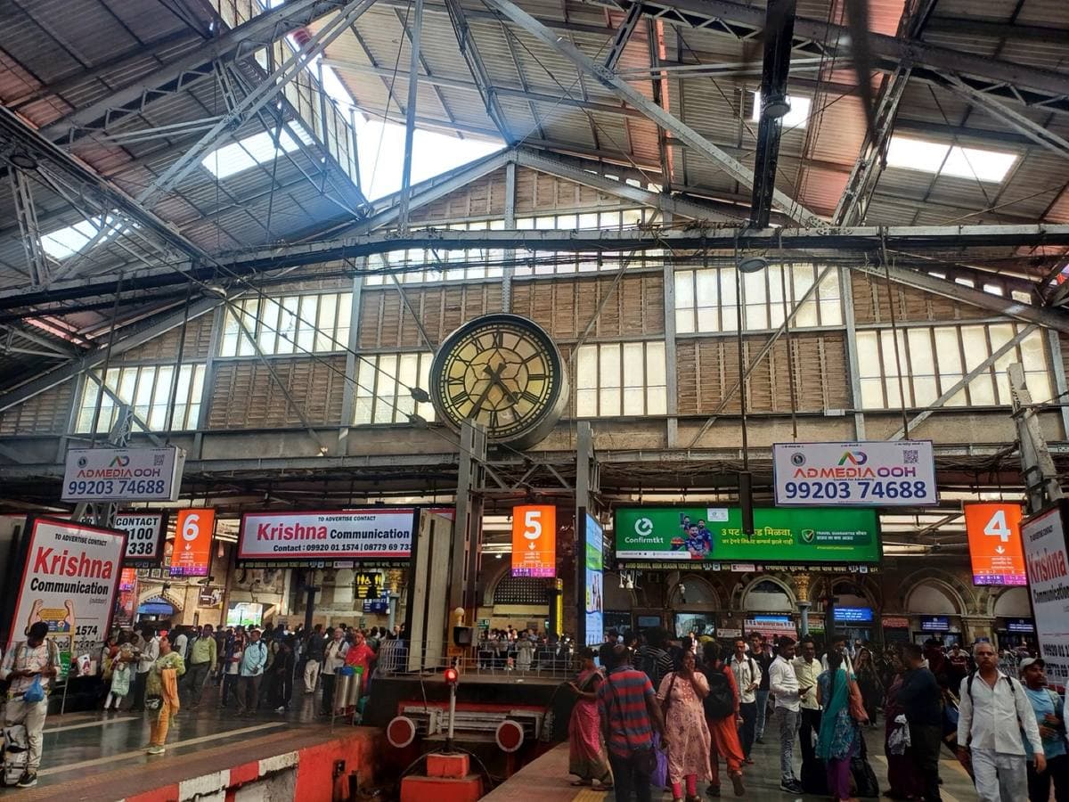 Colour photograph of a station concourse under a steel and glass roof, with a large clock, platform number signs and crowds of passengers