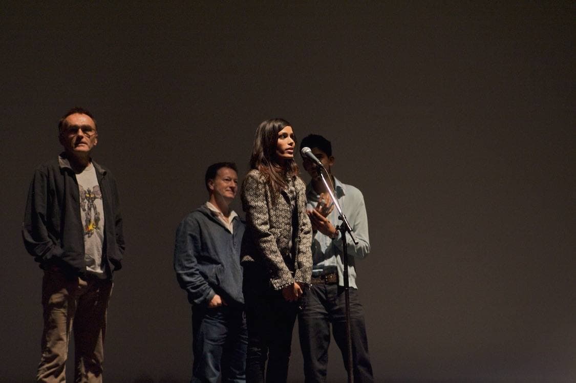 Colour photograph of four people on a dark stage, a young woman speaking into a microphone
