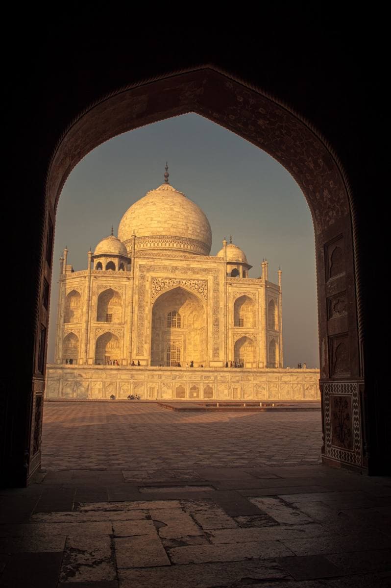 Colour photograph of the white marble Taj Mahal glowing orange at sunrise, seen through a dark sandstone arch