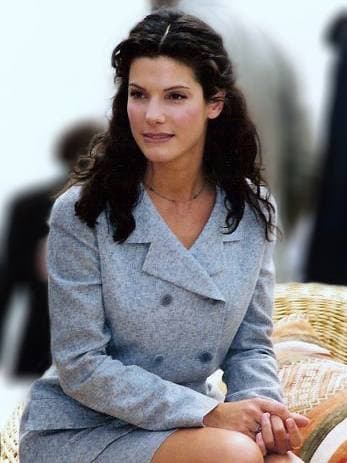 Sandra Bullock with long dark hair in a pale grey suit, seated